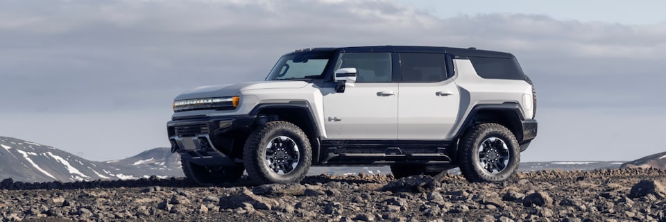 Side view of a white 2025 GMC Hummer EV SUV on a rocky plain under a beautiful blue sky.