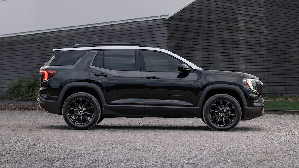 Vue latérale du GMC Terrain 2025 stationné seul devant une maison en bois sous un ciel gris.