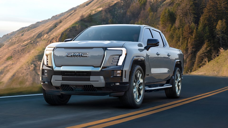 Vue latérale avant du GMC Sierra EV Denali 2025 en mouvement sur une route à flanc de colline.