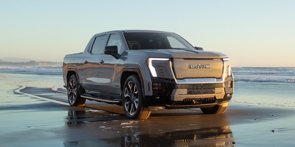 Vue latérale avant du GMC Sierra EV Denali 2025 stationné à la plage avec les phares allumés.