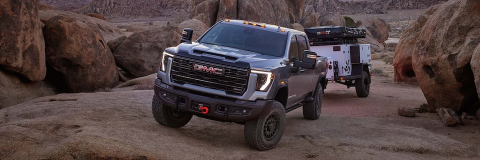 Front-side view of a 2025 GMC Sierra HD AT4 truck hauling a trailer through a rocky terrain at dusk.