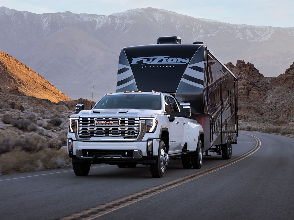 Un camion GMC Sierra HD Pro/SLE/SLT 2025 tire une grande remorque seul sur une route de montagne.