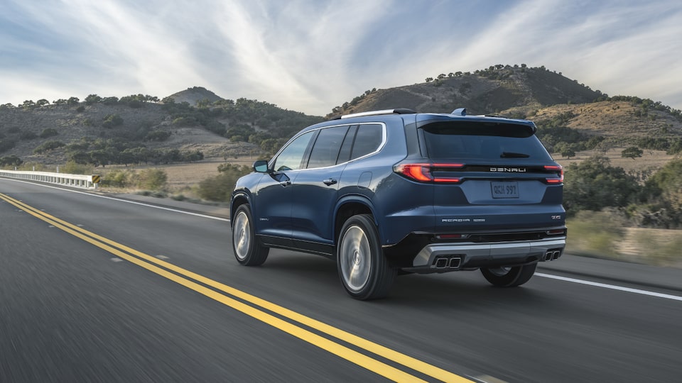 Back-side view of the 2025 GMC Acadia Denali driving on the road on a sunny day.