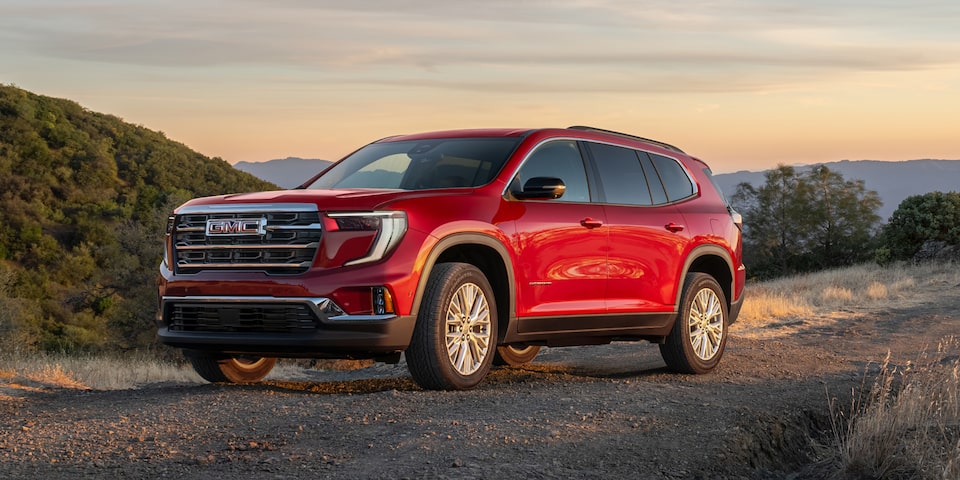 Front-side view of the 2025 GMC Acadia Elevation driving alone on a mountainside road at sunset.