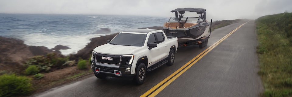 Front-side view of a 2026 GMC Sierra EV Pickup Truck towing a boat on a seaside road.