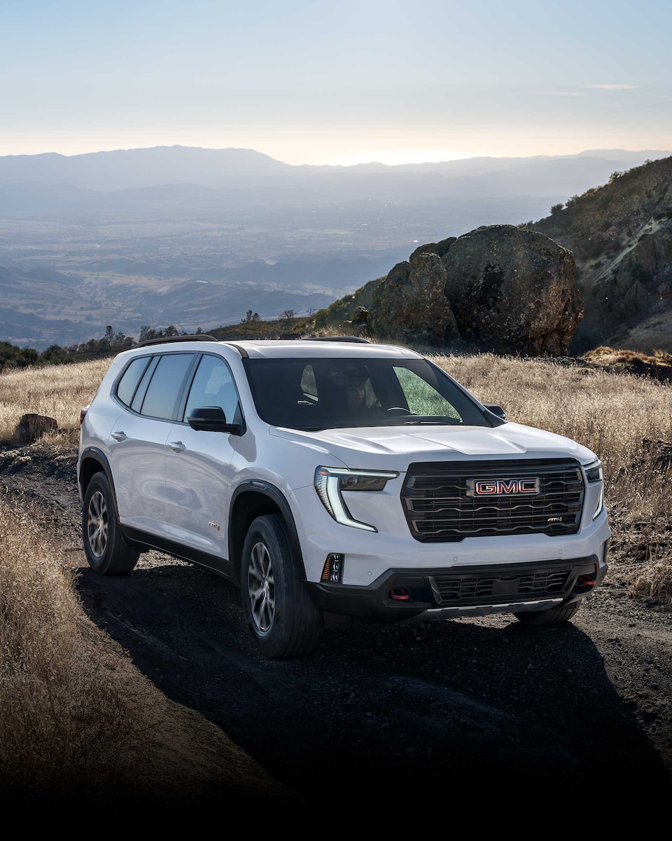 Front Three-Quarters View of a 2024 GMC Mid-Size SUV Driving on a Dirt Path with Mountains in the Background